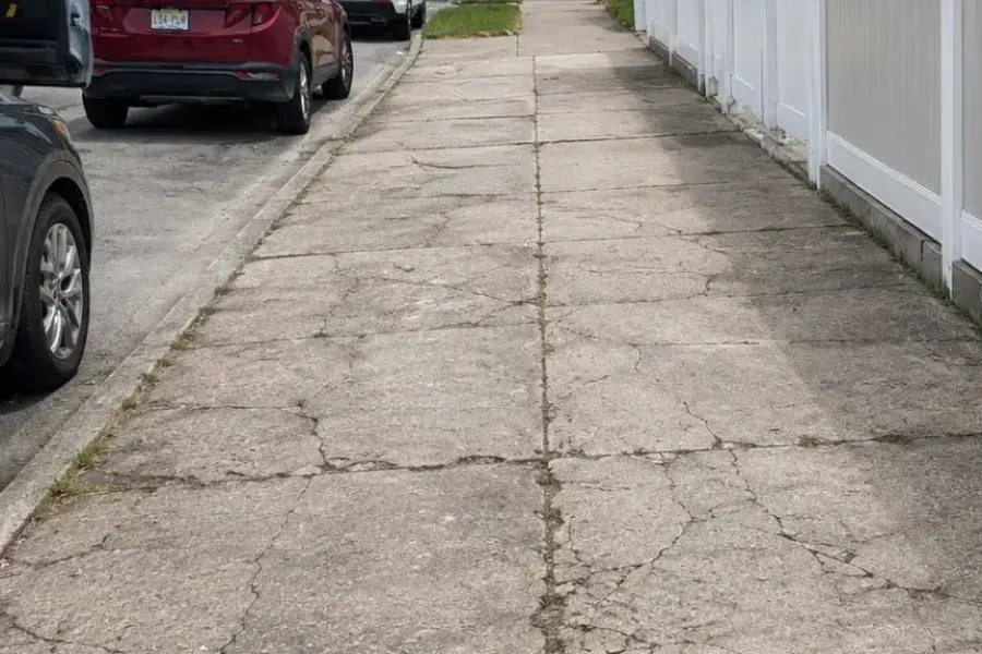 Before: Cracked and settled sidewalk flags beside a white fence in Queens before repair