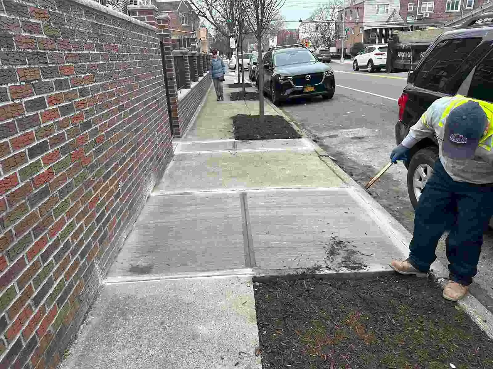 Worker finishing new sidewalk flags beside a brick wall and a tree pit, concrete work by Sidewalk Repair Near Me in NYC