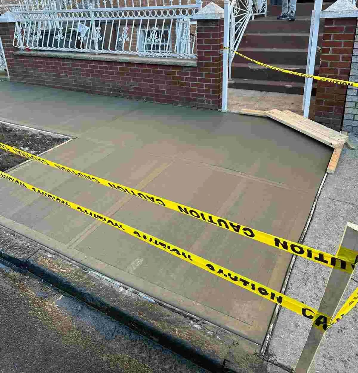 Fresh concrete sidewalk taped off beside a brick wall and a white iron fence, sidewalk repair by Sidewalk Repair Near Me in NYC